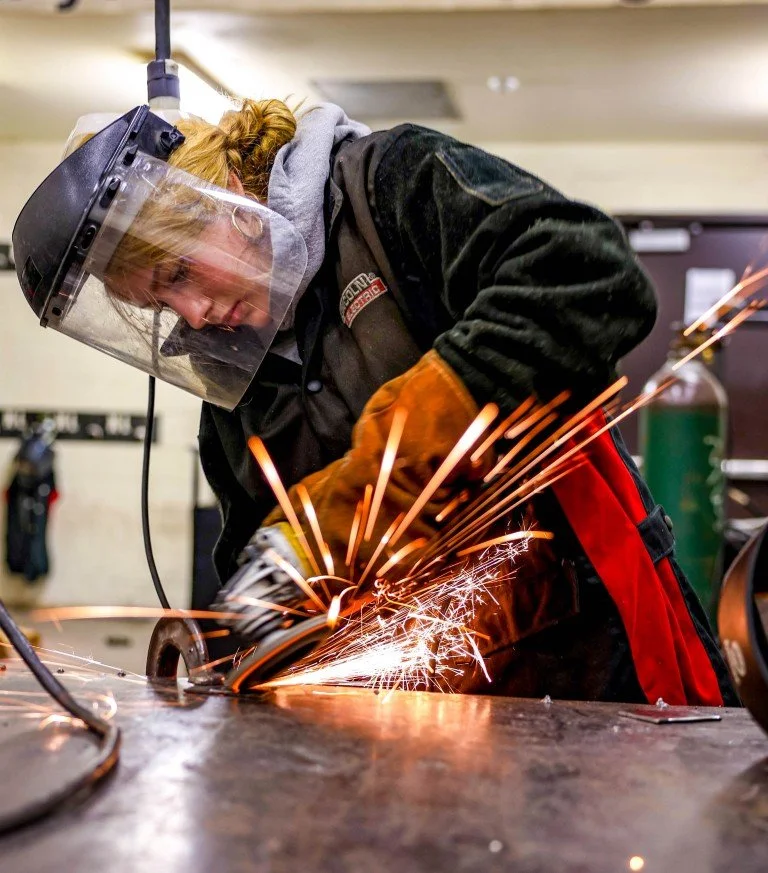 A person wearing a welding helmet, gloves, black jacket, and red apron is welding metal, producing bright sparks in a workshop.