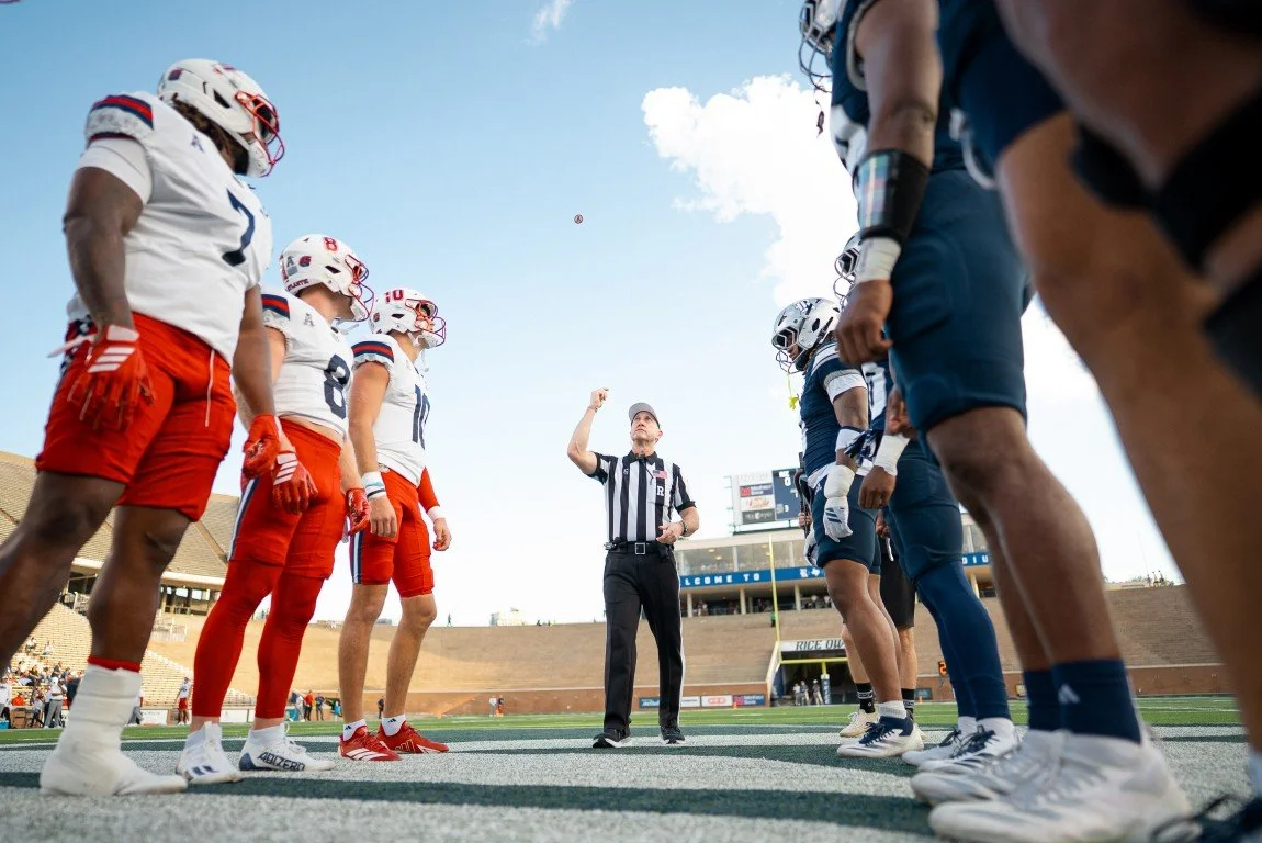 American football referees toss a coin at midfield before the game, with players from opposing teams in line for kickoff.