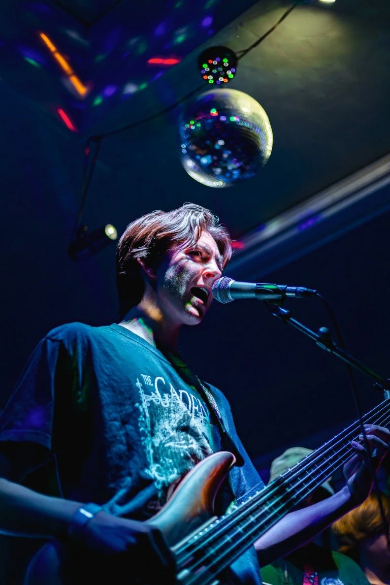 A young man singing into a microphone while playing an electric bass guitar on stage at a party or concert, with a disco ball and colorful lights overhead.