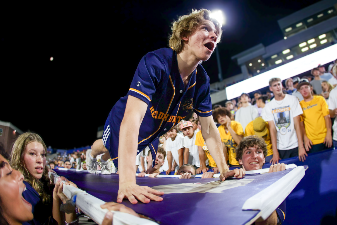 A young man in a navy blue sports jersey pushing a folding table during a sports event at night, surrounded by a crowd of young spectators wearing yellow and white shirts.