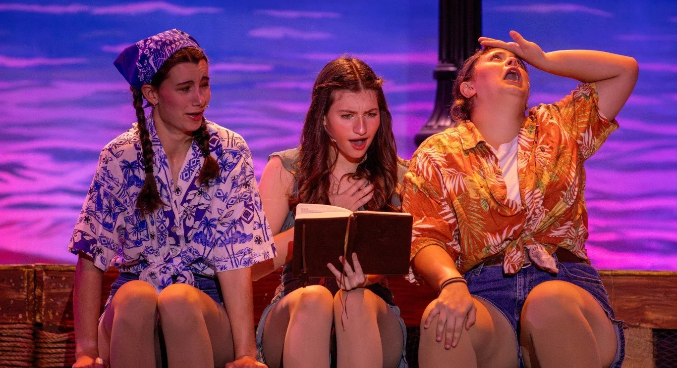 Three women sitting on a dock by the water, appearing distressed or emotional, with a purple and pink sunset background.