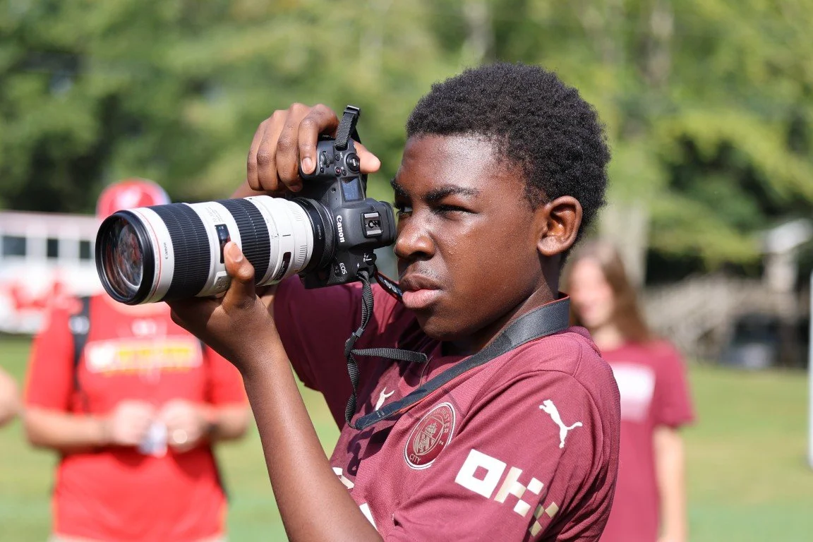A young man with short curly hair wearing a maroon sports jersey with PUMA logo, taking a photo with a Canon camera equipped with a large telephoto lens in an outdoor setting with green trees.