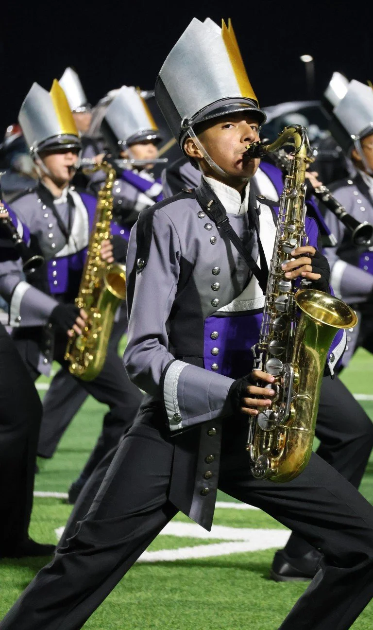 Band members in marching uniforms playing saxophones on a football field at night.