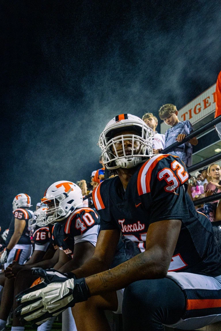 High school football players in black and orange uniforms sitting on the sidelines during a game, with spectators in the stands.