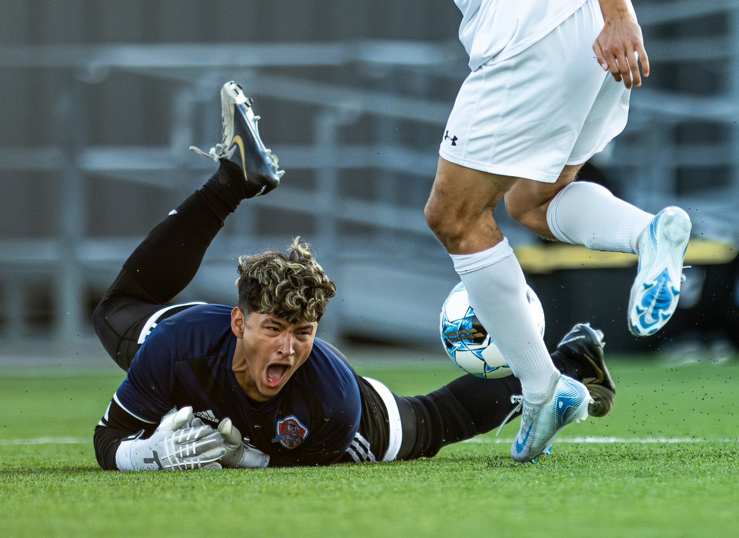 A soccer player in a white uniform leaps over a goalkeeper in a black uniform lying on the grass, during a soccer match.