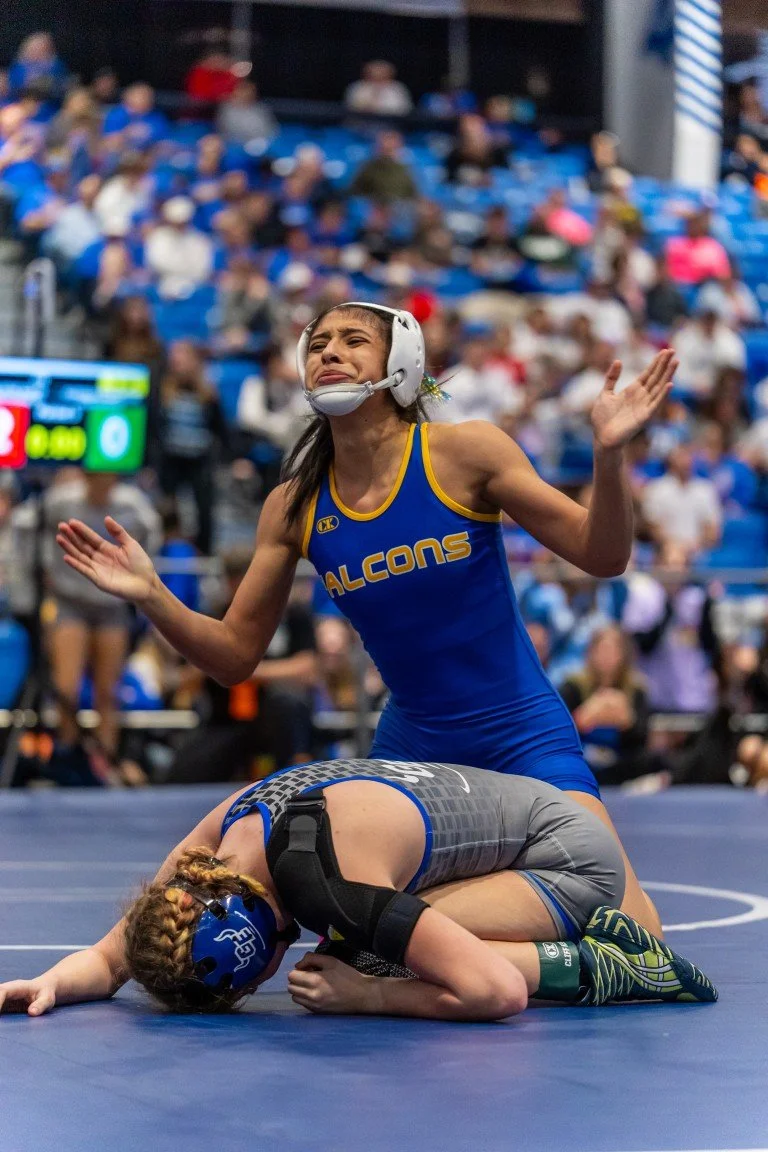 A female wrestler in a blue singlet and helmet is celebrating after winning, kneeling with arms raised in victory. A competitor in gray and blue is on the mat, head down and face turned away, appearing defeated. The background shows a crowded indoor 
