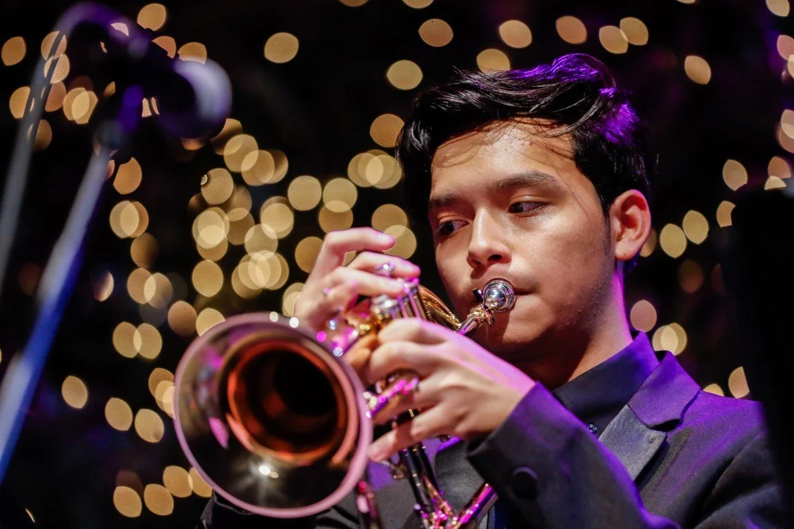 A young man in a black suit playing a trumpet during a performance, with blurred string lights in the background.