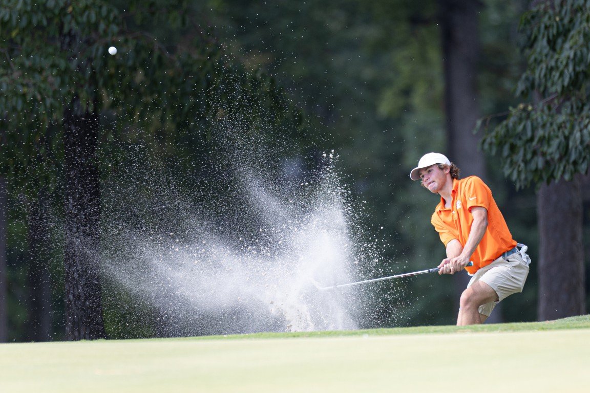 A young male golfer hitting a shot out of a sand trap on a golf course, with sand flying into the air.