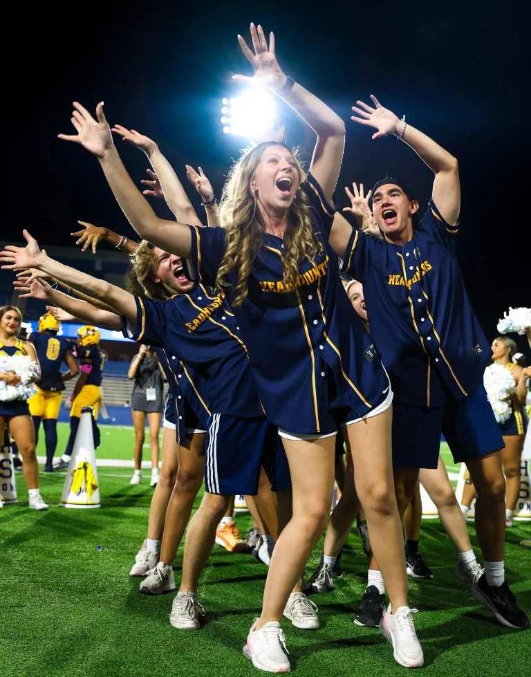 A group of young sports fans in matching navy blue jerseys with yellow accents celebrating on a football field at night, arms raised, smiling, with stadium lights in the background.