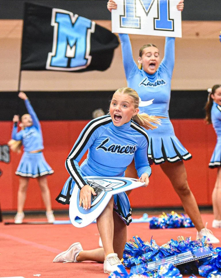 Cheerleaders dressed in blue cheer on their team during a game, with some holding signs and flags and others cheering energetically on the court.