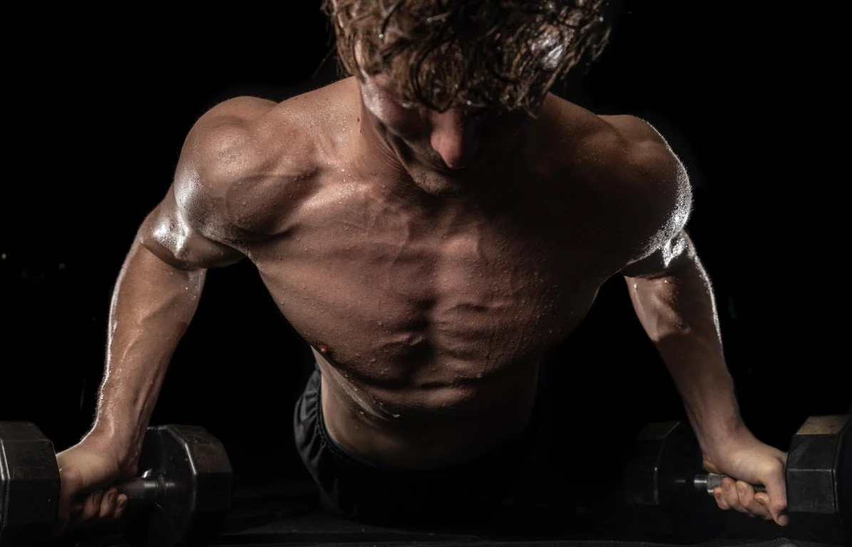 A shirtless man with wet hair and a muscular build performing a push-up with dumbbells, surrounded by a black background.