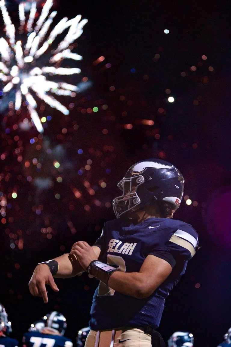 An American football player in a dark blue uniform and helmet stands under fireworks at night.