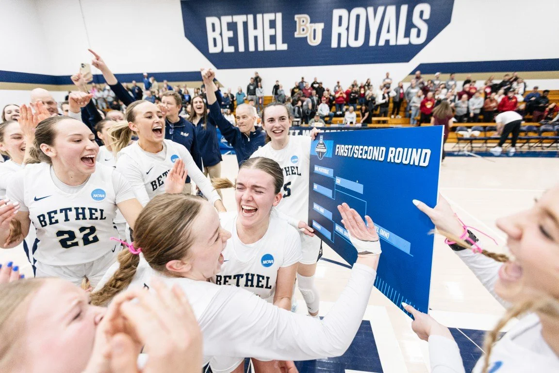 Bethel women's volleyball team celebrating with their coach after winning a match, holding a bracket sign on the court, with spectators in the background at Bethel University.