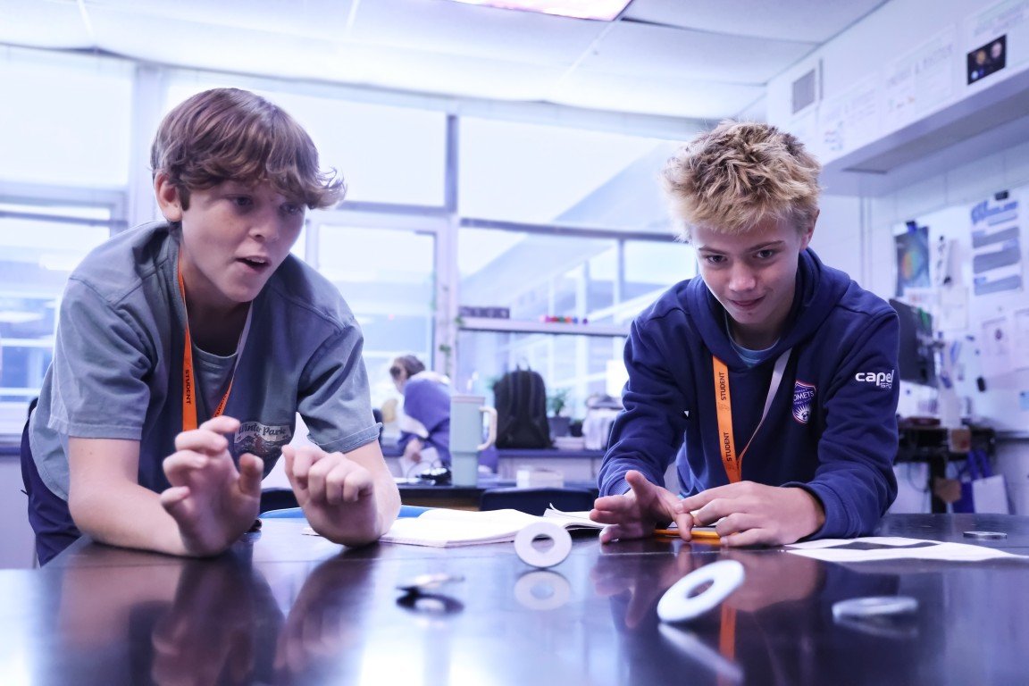 Two boys in a science classroom focusing on an experiment or activity involving some circular objects on the table.