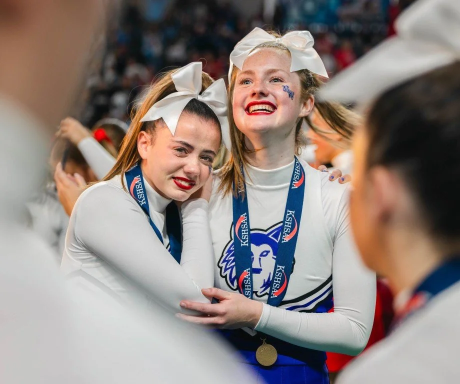 Two young female cheerleaders with white bows in their hair and white uniforms, celebrating at a sports event, wearing medals.