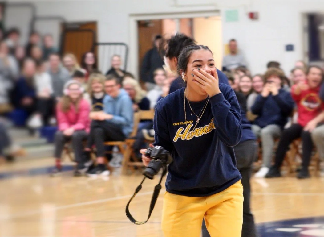 A young woman with dark hair tied back, wearing a navy sweatshirt with 'Kirtland Hornets' written on it and yellow pants, is holding a camera and laughing with her hand covering her mouth. She is in a school gymnasium in front of a cheering crowd of 