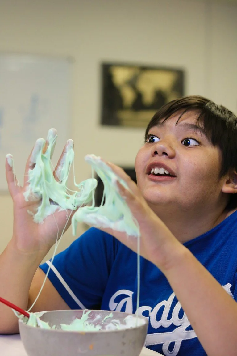 A boy with dark hair and a blue shirt has green slime on his hands, stretching it between his fingers with a surprised expression.
