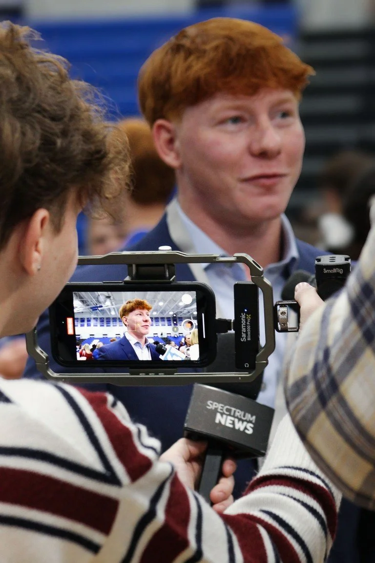 A young man with red hair in a blue suit being interviewed by the media, with a microphone labeled 'Spectrum News' and a camera capturing his image at an indoor event.