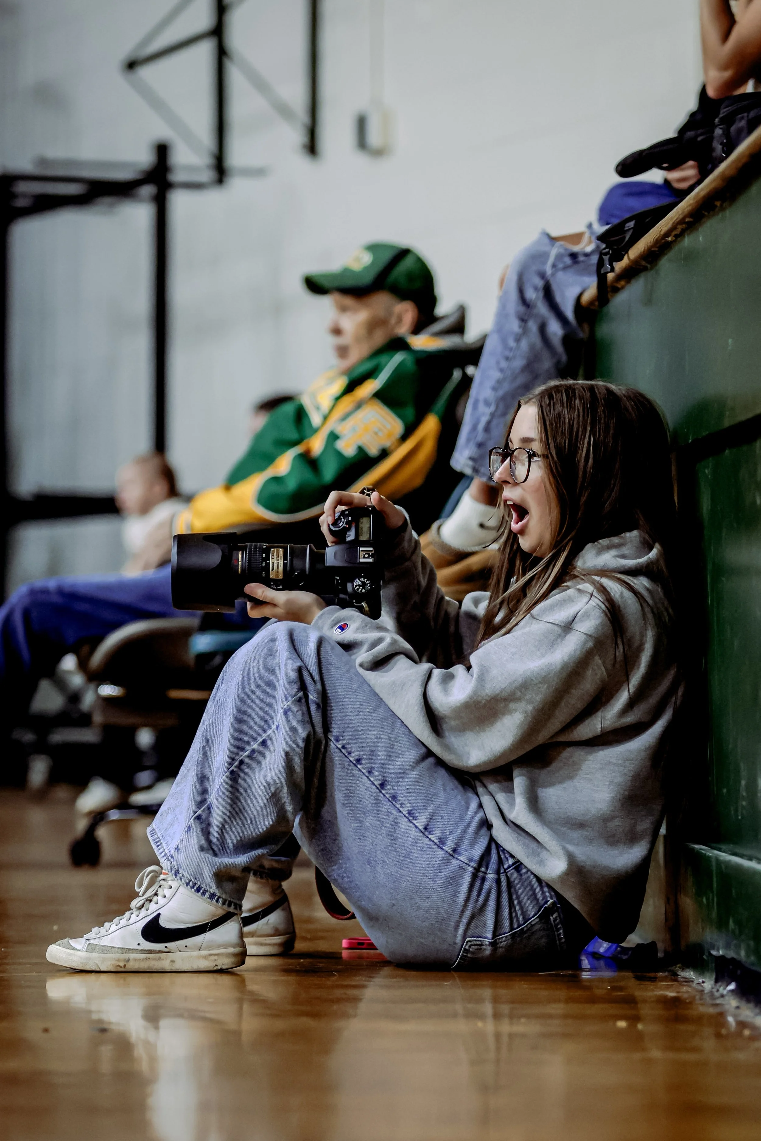 A young woman with glasses and long hair sitting on the floor of a gymnasium, holding a camera and looking excited or surprised. She is wearing a gray sweatshirt, baggy jeans, and white Nike sneakers with black swoosh. In the background, there are ot
