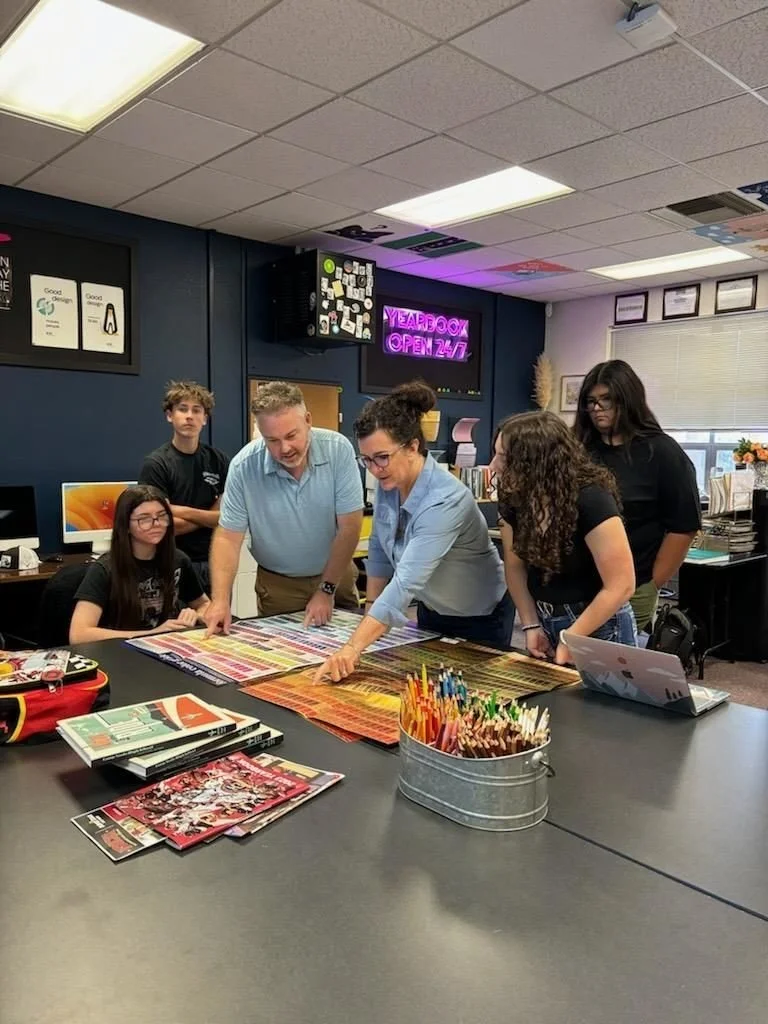 Group of six people, including two adults and four young adults, gathered around a table in an office, looking at colorful fabric or sample swatches. The office has a dark blue wall with framed certificates, a neon pink sign that says 'Yearbook Open 