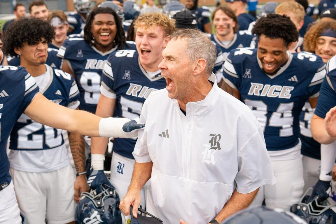 Football coach in white jacket passionately addressing his team, who are wearing navy blue and white uniforms, during a game or rally. The team is smiling and appears enthusiastic.