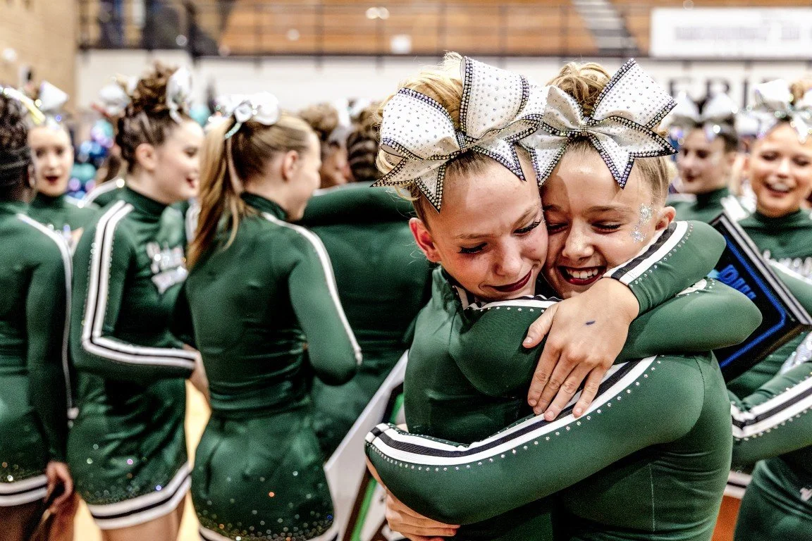 Two young cheerleaders in green uniforms with white and black accents hugging each other in celebration, surrounded by other cheerleaders in the background.