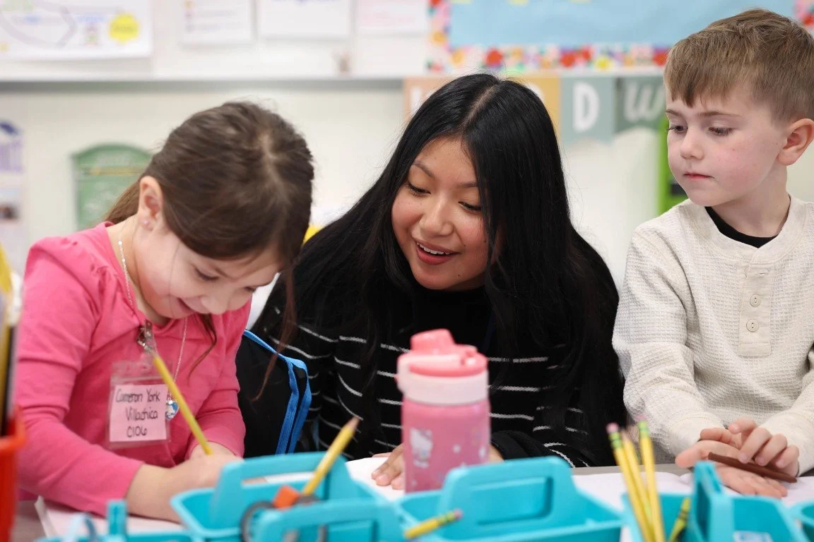 A young girl and a woman are smiling while working together on a school activity, with a boy next to them observing. They are in a classroom setting with educational posters and supplies on the table.