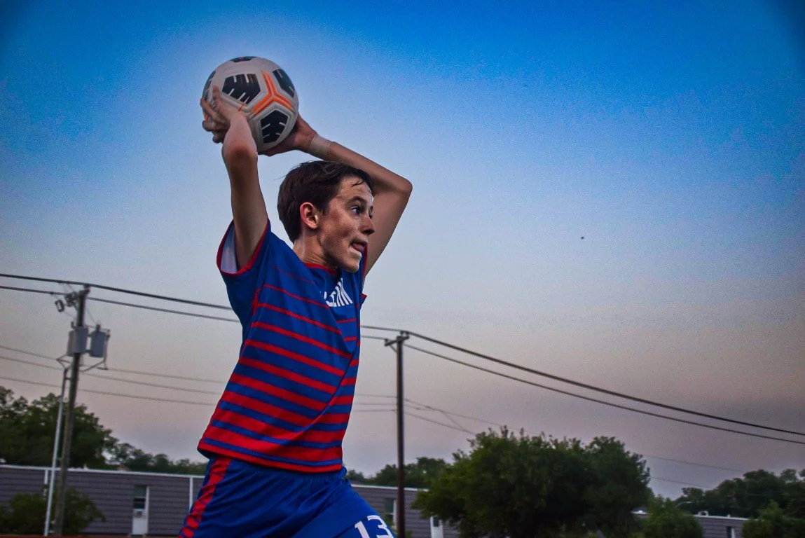 A young male soccer player in a blue and red striped uniform preparing to throw a soccer ball during sunset outdoors.