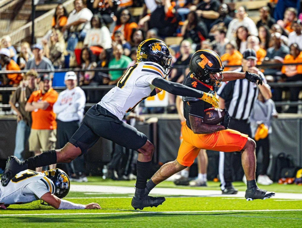 A football game action shot showing a player in a black and orange uniform running with the football while being chased by a player in a white and black uniform. The players are on a green field with spectators in the stands watching.