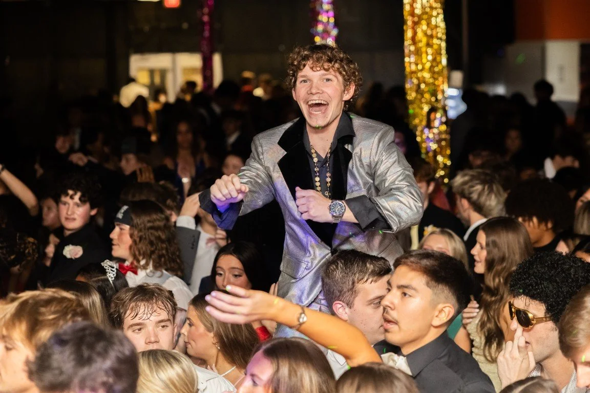Young man with curly hair, wearing a shiny silver blazer, smiling and raising his arm at a crowded dance party or celebration.