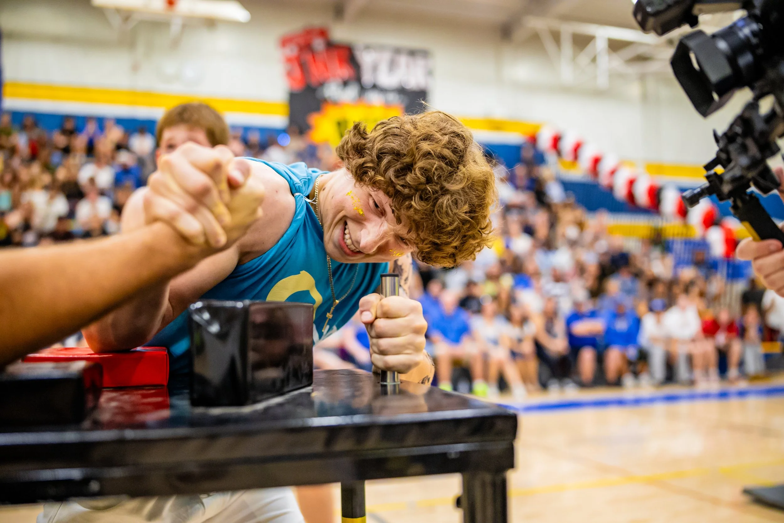A young man with curly hair and a blue shirt participates in a strength competition, gripping an arm wrestling machine's handles and smiling. The background shows a cheering crowd in a gymnasium.