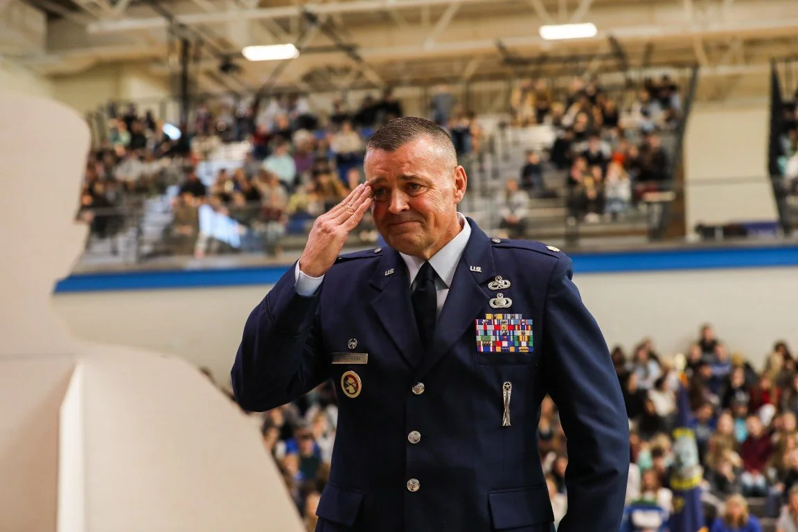 A man in U.S. Air Force uniform saluting in front of a large crowd at an indoor event.