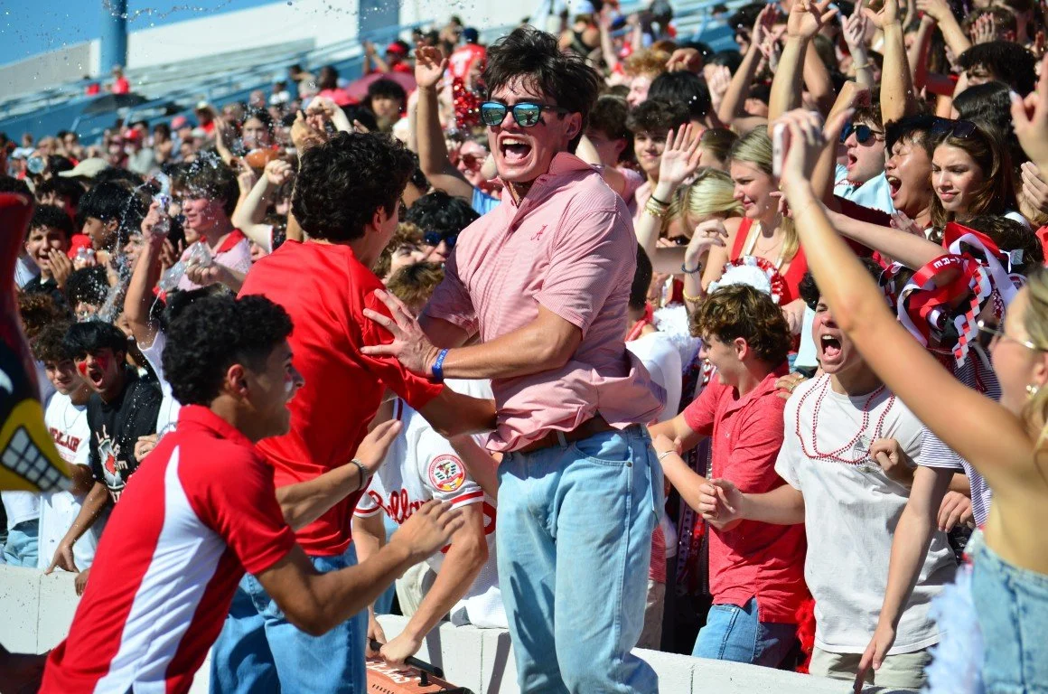 Crowd of excited sports fans at a stadium, celebrating and cheering, with a focus on a man wearing sunglasses and a pink shirt, erupting in joy with others around him.