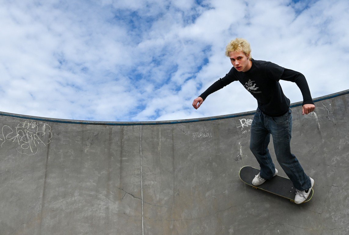Young man with blonde hair skating on the edge of a concrete skate park bowl under a partly cloudy sky.