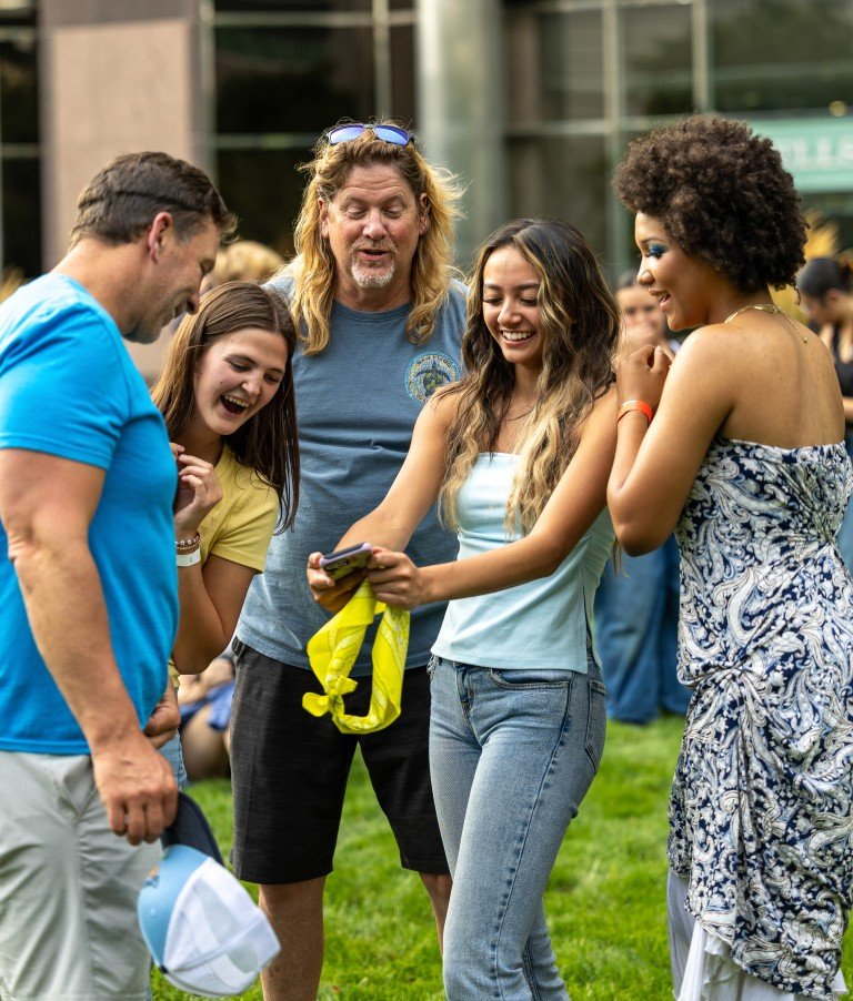 Group of five people standing outdoors, looking at a phone and smiling, in a casual, happy gathering.