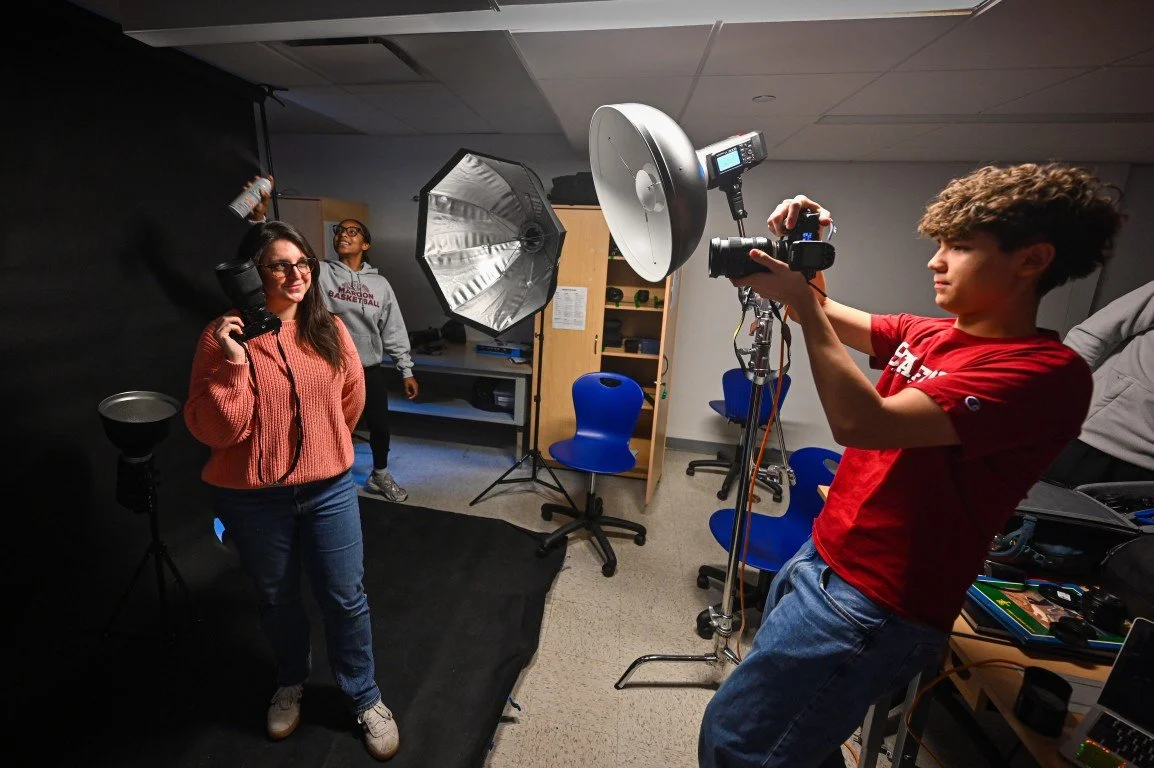 Interior of a photography studio with two women and a young man. One woman is adjusting her glasses and holding a camera with a softbox light, while the other woman in the background is standing near a large umbrella light. The young man, wearing a r