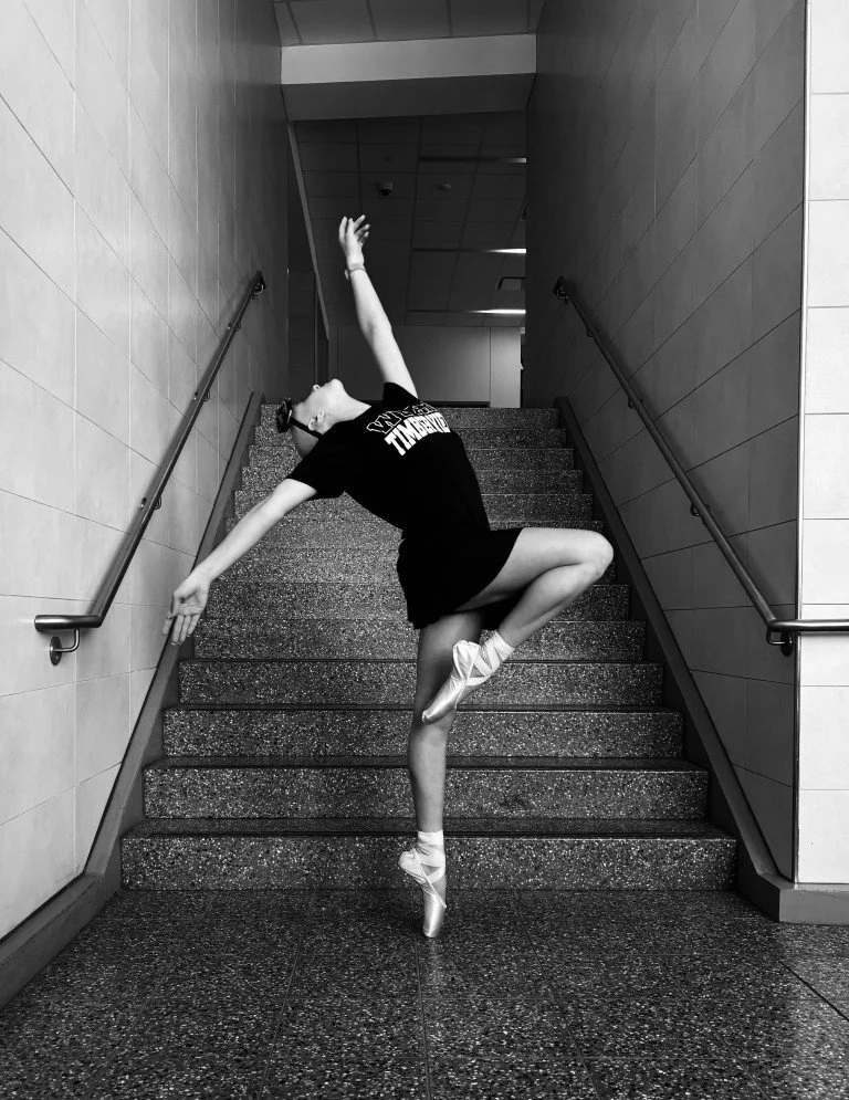 A ballet dancer practicing on a staircase indoors, wearing ballet slippers, a black T-shirt, and shorts, striking a graceful pose.