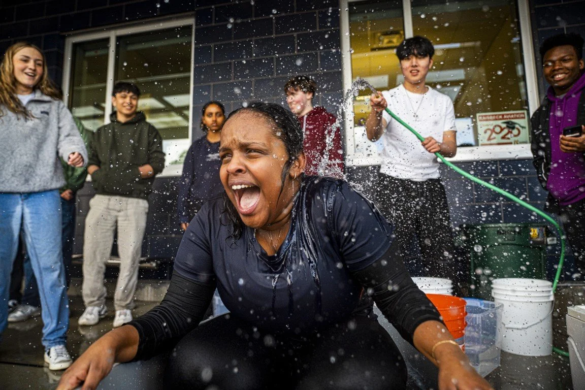 A woman in a black shirt is soaking wet and cheering while a group of smiling young people watch her outside a building, with one person spraying her with a hose.