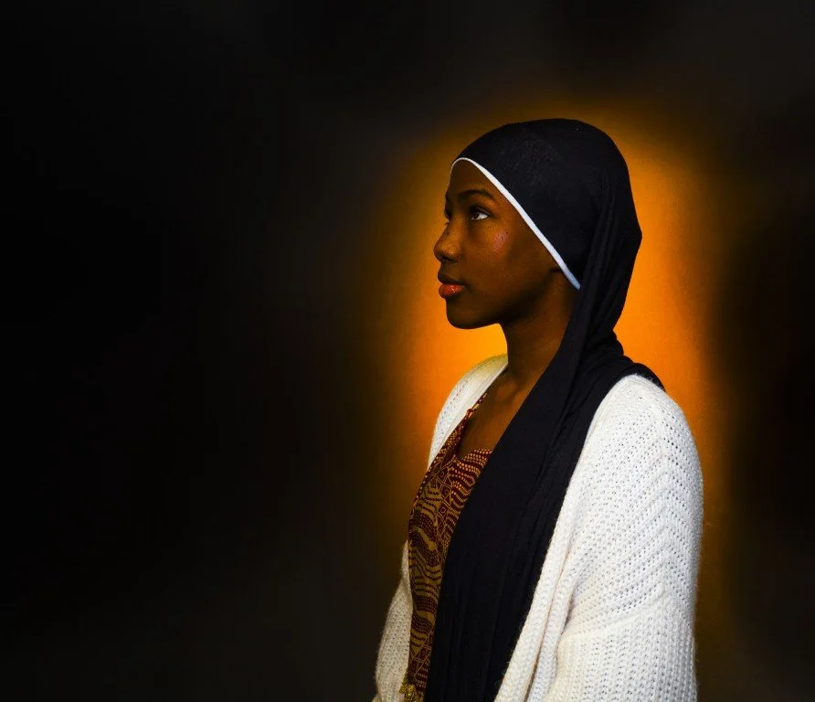 Portrait of a woman in profile with a black headscarf and white sweater, standing against a dark background with warm orange backlighting.