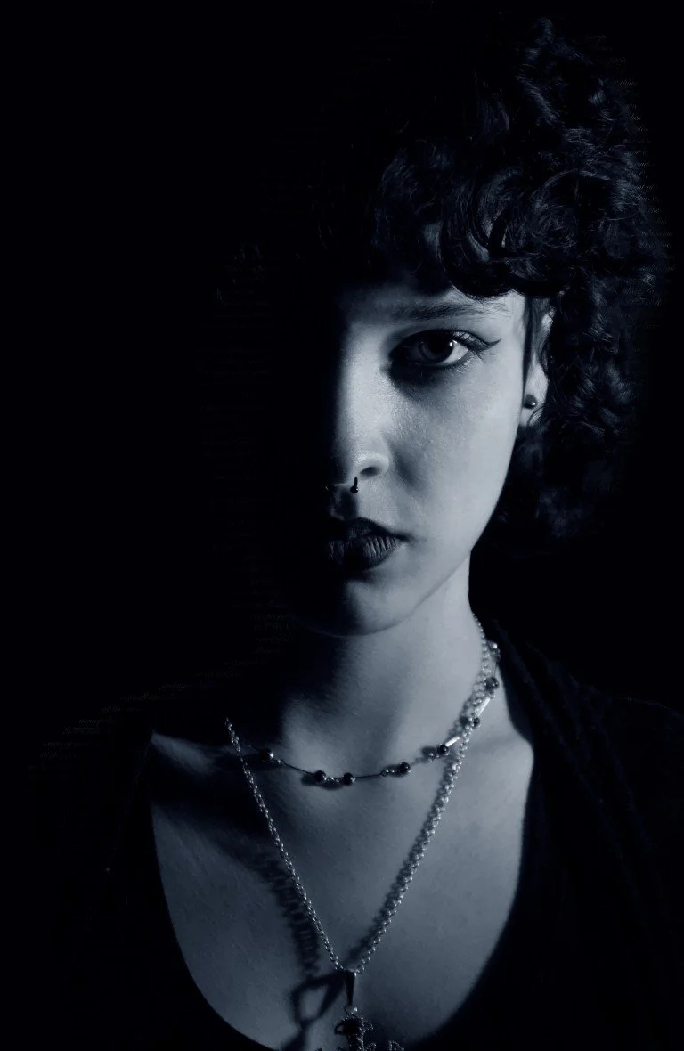 A woman with curly hair and multiple necklaces posing in low light, with half of her face illuminated and the other half in shadow.