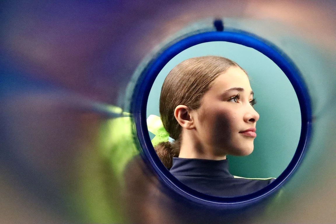 A young woman with brown hair tied back, viewed through the opening of a sports bottle, smiling gently with a neutral background.