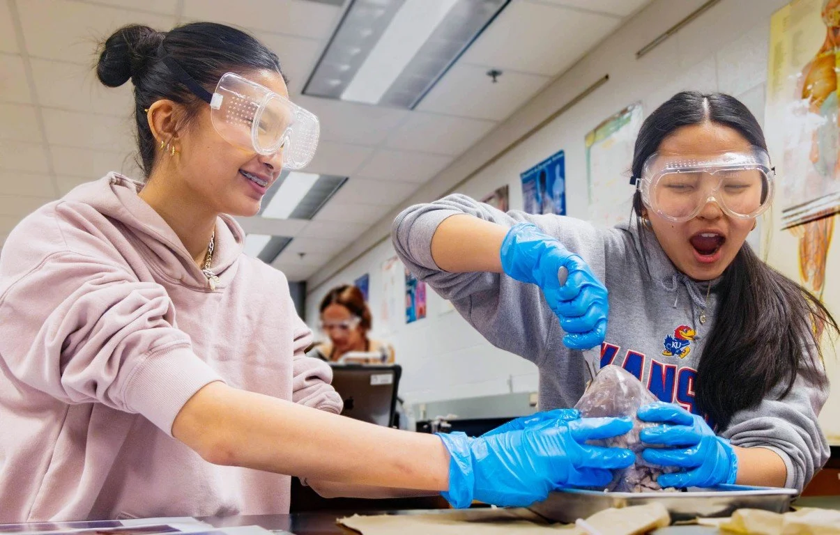Two women wearing safety goggles and gloves, handling an ice block in a classroom or laboratory setting, with posters on the wall.