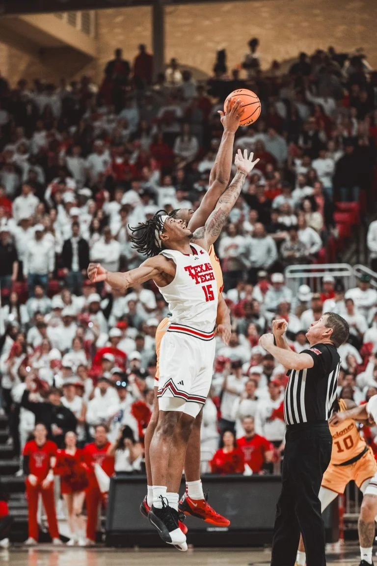 A basketball game scene with a player from Texas Tech jumping to shoot or block a shot, while a referee watches. Spectators fill the background.