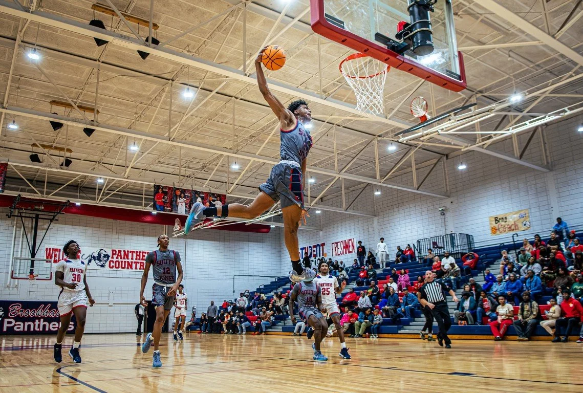 A basketball player is mid-air attempting a dunk during a game, with players and spectators watching. The gym has a high ceiling, scoreboard, and crowd seated on bleachers.