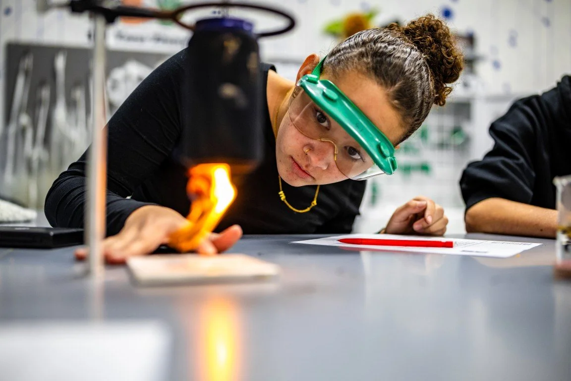 A woman wearing safety goggles and a green headband observes a flame from a Bunsen burner in a science laboratory.