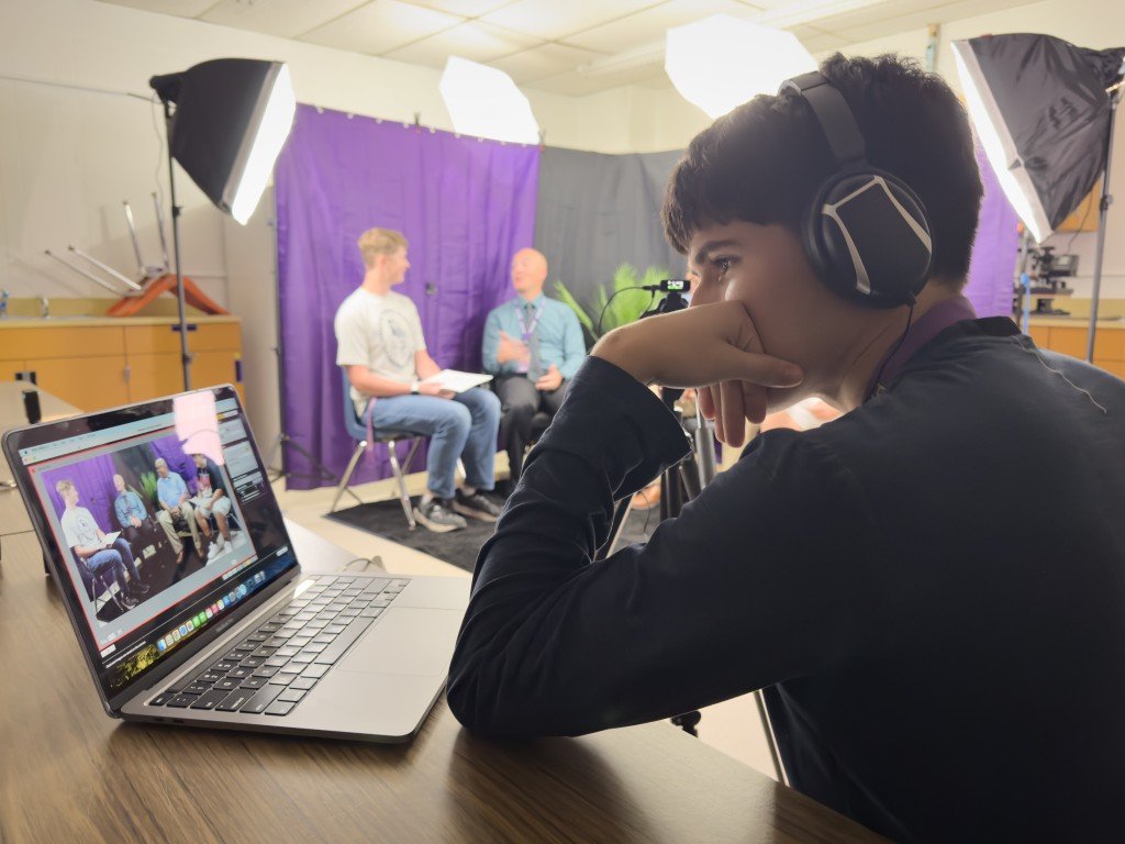 Person wearing headphones operating a laptop during a video recording session with two people in the background on a set with purple curtains.