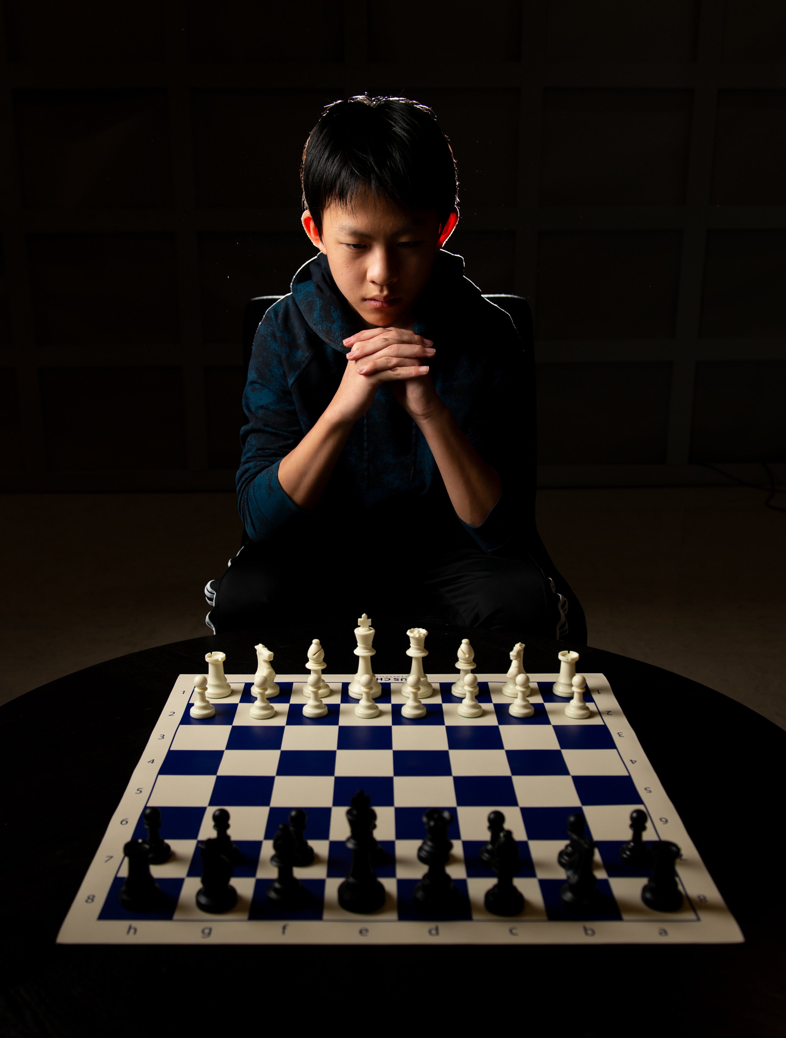 A young male chess player with black hair, wearing a blue hoodie, sitting at a chessboard with pieces in starting position, contemplating his move. The photo has dramatic lighting and a dark background.