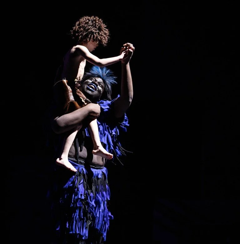Performer with face painted black and blue costume holding a child with curly hair on their shoulders, with a dark background.