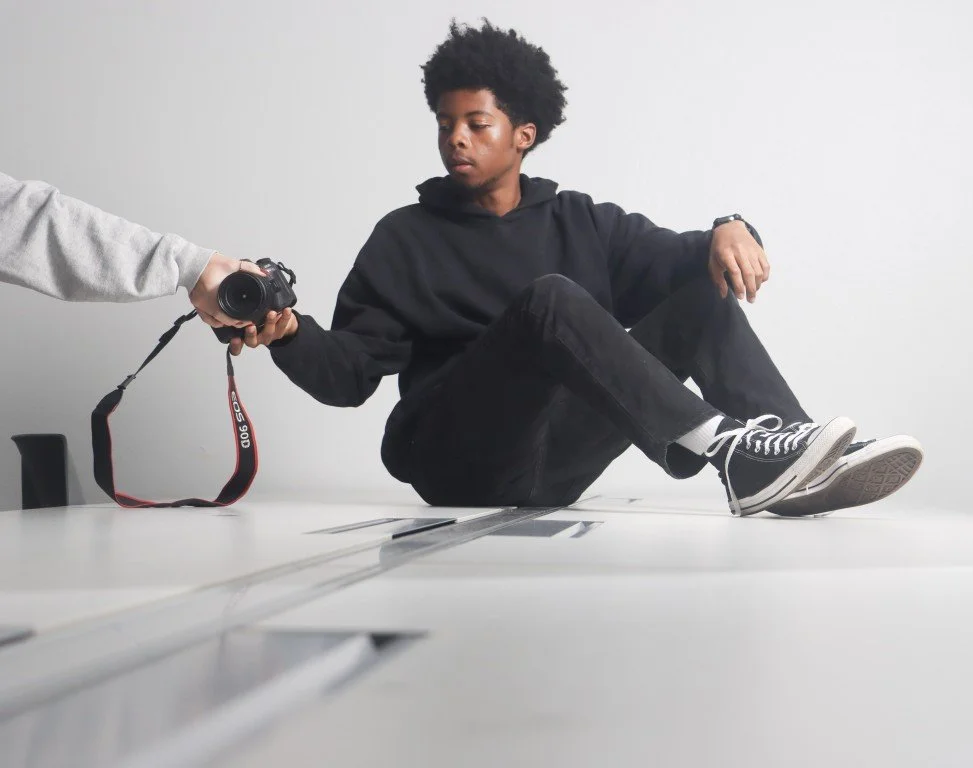 Young man with curly hair sitting on a white table, holding a camera being handed to him by another person out of view. The young man is wearing a black hoodie, black pants, and black sneakers with white soles and laces.