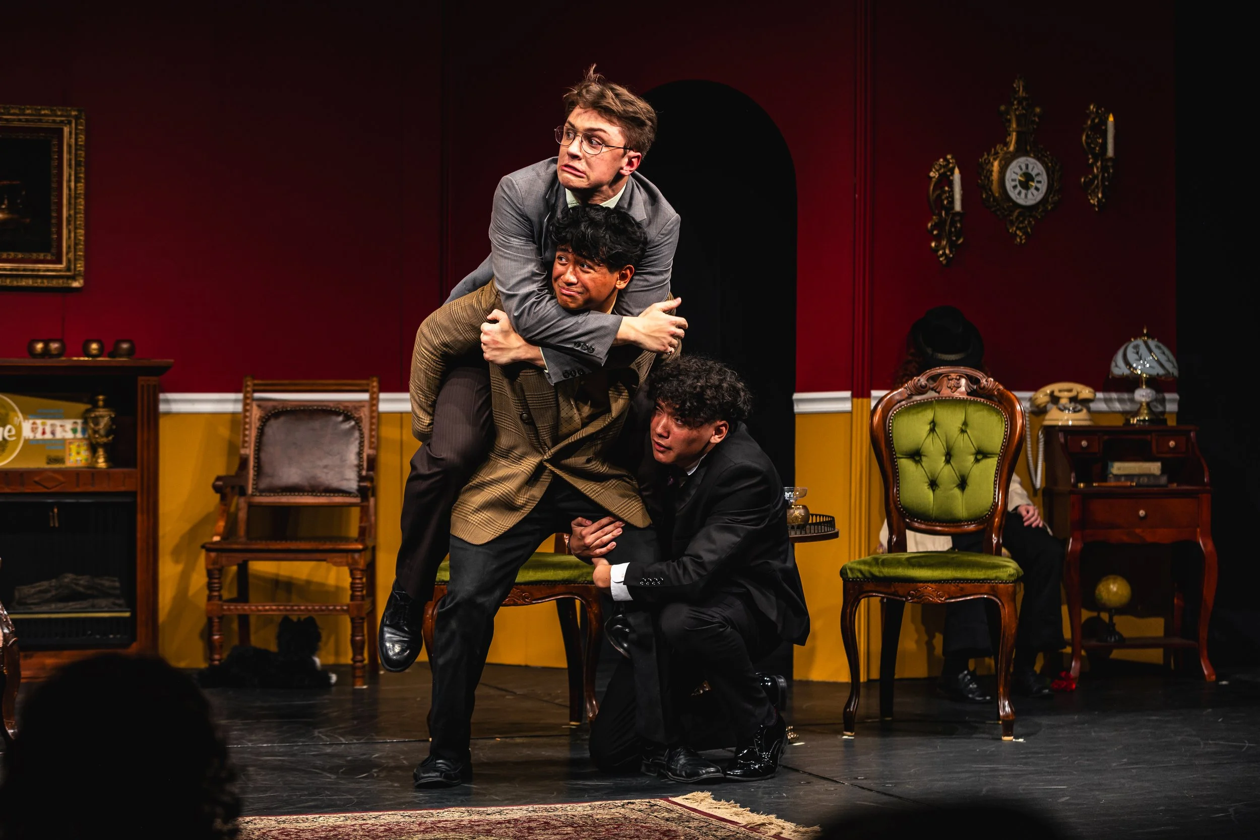 Four men performing a comedy skit on stage, with one man sitting on a chair and the others standing and leaning on him, in a room decorated with antique furniture and wall clocks.